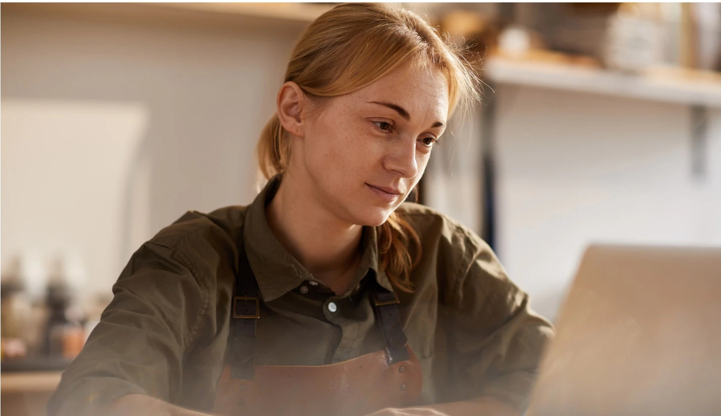 Woman in a crafting apron working on a laptop in a workshop at sunset